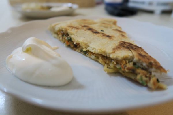 פרוסת לחם שטוח ממולא על צלחת לבנה, מלווה בכוס שמנת חמוצה. נראה שמלית הלחם השטוח כוללת ירקות ואולי בשר. הצלחת מונחת על משטח בהיר.