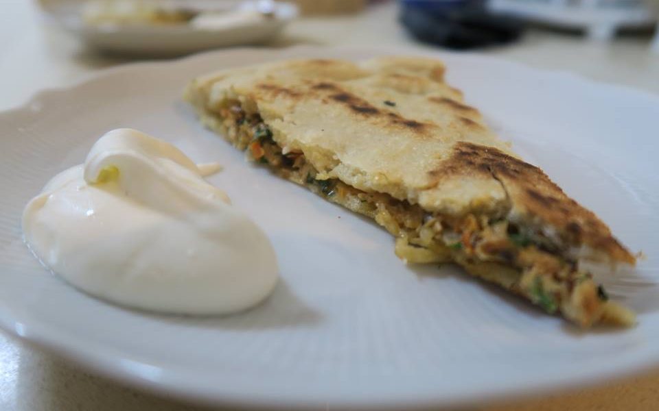 פרוסת לחם שטוח ממולא על צלחת לבנה, מלווה בכוס שמנת חמוצה. נראה שמלית הלחם השטוח כוללת ירקות ואולי בשר. הצלחת מונחת על משטח בהיר.