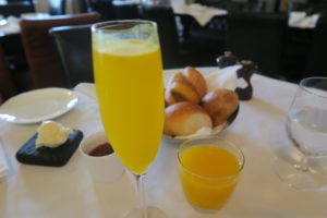 כוס מיץ תפוזים וכוס גבוהה של מימוזה מונחות על השולחן. מאחוריהם יש סלסלת לחמניות, צלחת קטנה עם חמאה, מנה נוספת עם רוטב אדום, כוס מים ושייקרות מלח ופלפל על מפה לבנה.