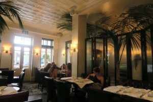 A group of people sit at a dining table in a restaurant with tall palm plants and elegant decor. Sunlight filters through large windows, illuminating the room. Tables are set with white cloths and crockery, and chairs are upholstered in dark material.