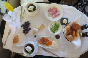 A table set for brunch with a plate of smoked salmon, bowls of poached eggs, a variety of breads, a slice of cheese, sliced ham, grapes, sliced cucumbers, a glass of orange juice, and butter. There are salt and pepper shakers on the side.