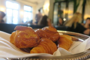 A silver tray with croissants is placed on a white napkin. The background shows an out-of-focus cafe interior with people sitting at tables.