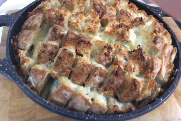 תבנית שחורה עגולה מחזיקה צלחת אפויה של לחם פרוס עם חלק עליון. חתיכות הלחם נחתכות לתבנית רשת ונראה שיש גבינה מותכת או תוספת דומה בין הנתחים. המחבת על משטח עץ.