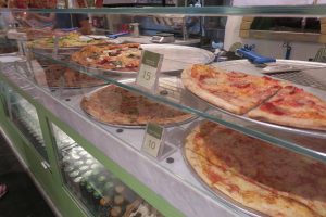A display case showing various slices of pizza on trays, including pepperoni and cheese varieties. Each slice has a price tag, with numbers 15 and 10. Canned drinks are visible in a cooler below the counter.