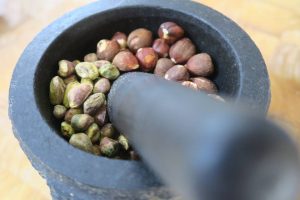 מכתש שחורה המכילה אגוזי לוז ופיסטוקים, ובתוכו נח עלי. את המכתש והעלי מניחים על משטח עץ. התמונה מתמקדת במבט העליון בתוך המרגמה.