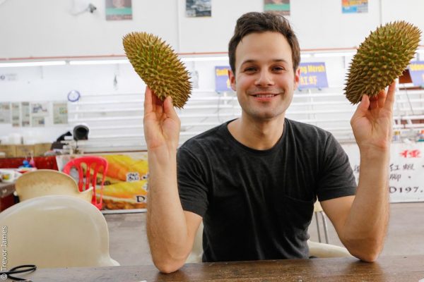 אדם עם שיער כהה קצר יושב ליד שולחן, מחייך, ומחזיק שני דוריאנים בידיו. ברקע נראים כיסאות ושולחנות ריקים עם כמה שלטים המציגים טקסט בשפה אסייתית.