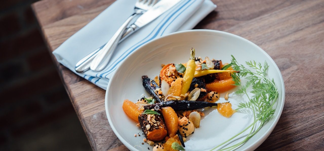 צלחת לבנה על שולחן עץ מחזיקה מנה עם גזר צלוי, קוביות קטנות של רוטב וקישוטים. מפית עם מזלג וסכין מונחת ליד הצלחת.