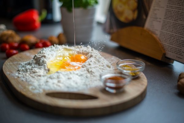 קרש חיתוך מעץ ובמרכזו תלולית קמח ובו ביצים סדוקות. בקרבת מקום יש קערות זכוכית עם תבלינים ושמן. ברקע יש ספר בישול, עגבניות, אגוזי מלך, פלפל אדום וצמח.