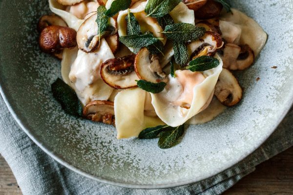 צלחת קרמית במילוי רביולי, בציפוי פטריות שחומות ועלי מרווה פריכים, על מפית אפורה במרקם. משטח השולחן עשוי עץ.