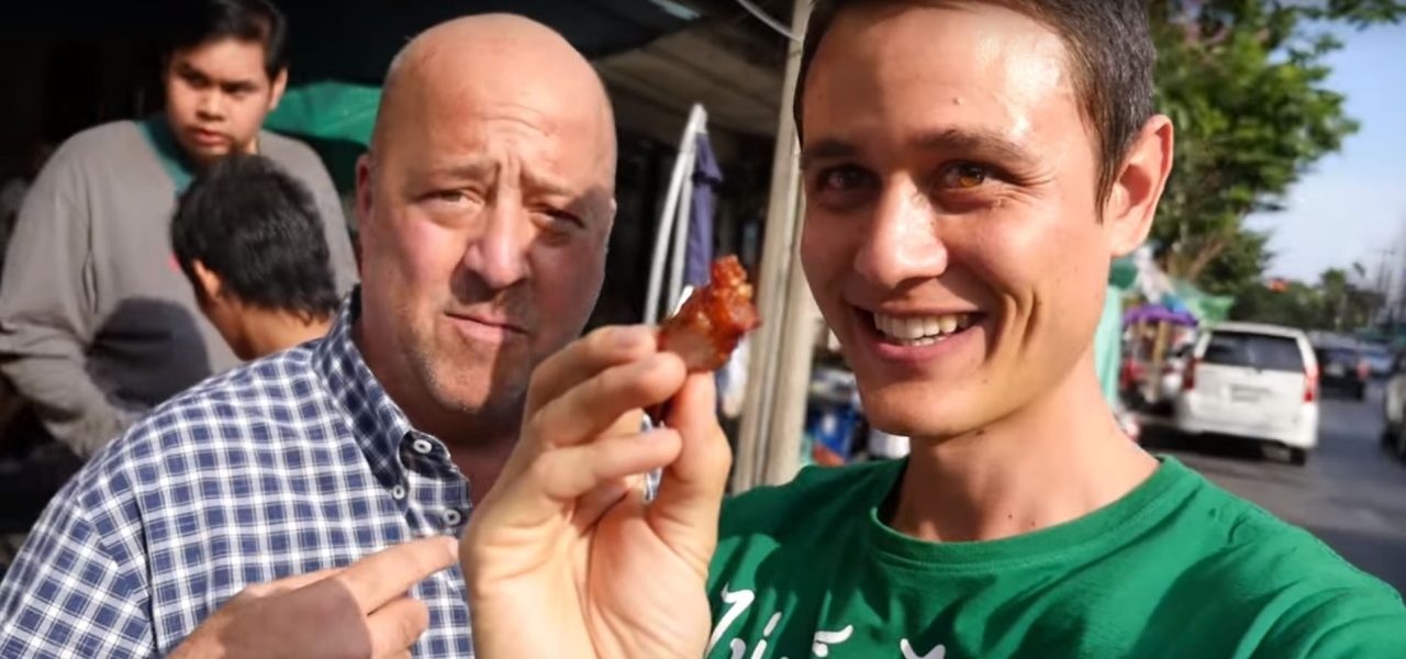 שני גברים עומדים בחוץ כשאחד מחזיק חתיכת אוכל קרוב למצלמה. האיש משמאל בחולצה משובצת מביט במצלמה, בעוד האיש מימין בחולצה ירוקה מחייך ומחזיק את האוכל.