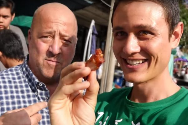 שני גברים עומדים בחוץ כשאחד מחזיק חתיכת אוכל קרוב למצלמה. האיש משמאל בחולצה משובצת מביט במצלמה, בעוד האיש מימין בחולצה ירוקה מחייך ומחזיק את האוכל.