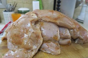 חזה עוף גולמי ומתובל מוערמים על קרש חיתוך מעץ בסביבת מטבח. פריטי מטבח שונים מטושטשים ברקע, ביניהם מיכל עם כלים וטחנת פלפל.