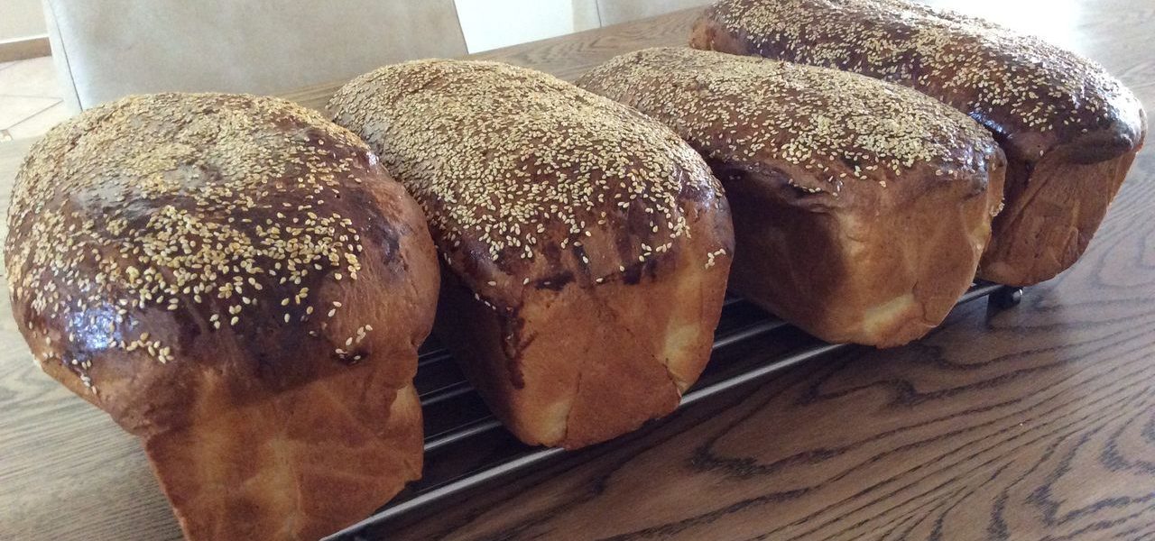 ארבע כיכרות לחם חום זהוב בציפוי שומשום מוצגות על מתכת קירור מתכת. הם מסודרים בשורה על שולחן עץ עם כיסאות בז' ברקע.
