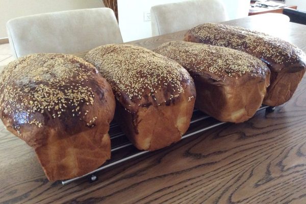 ארבע כיכרות לחם חום זהוב בציפוי שומשום מוצגות על מתכת קירור מתכת. הם מסודרים בשורה על שולחן עץ עם כיסאות בז' ברקע.