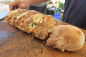 מספר פרוסות של לחם ממולה מסודרות באלכסון על קרש חיתוך מעץ. נראה שהלחם ממולה ברכיבים כמו גבינה וירקות. חלק מגופו של אדם נראה ברקע, שמחזיק את הקרש.