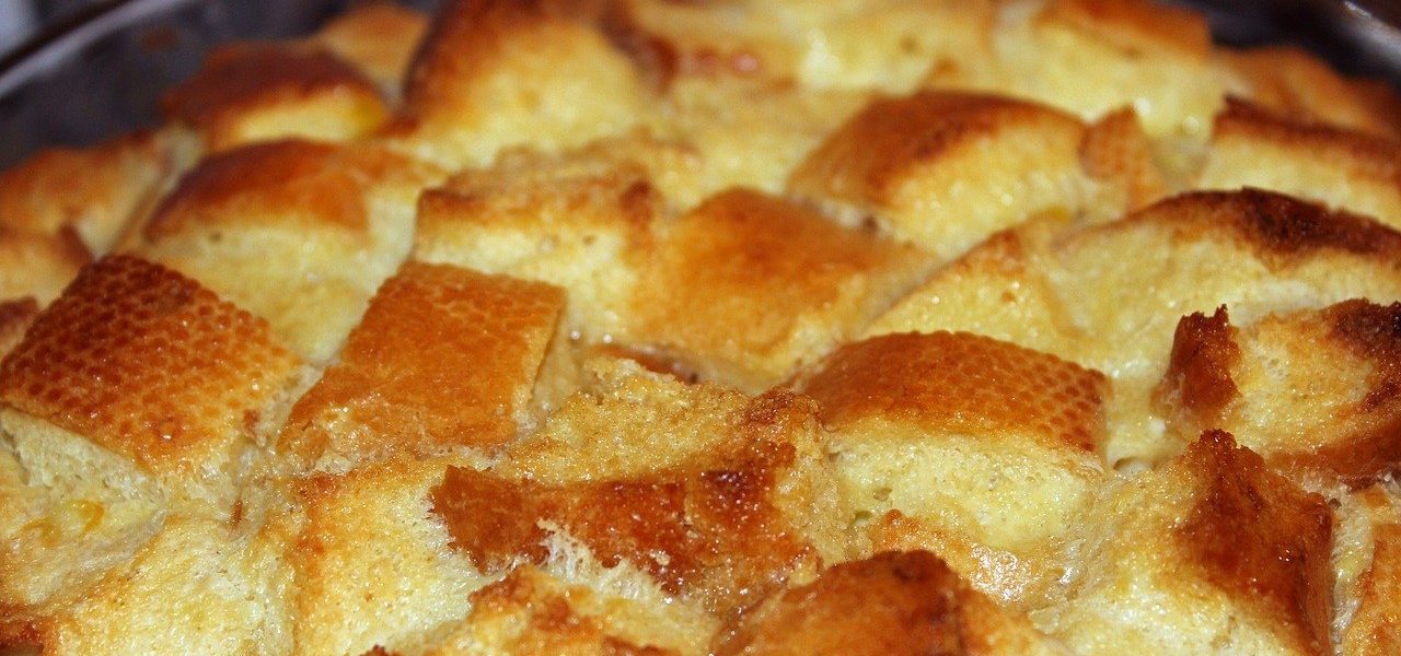 מבט מקרוב על תבנית אפייה מלאה בפודינג לחם חום-זהוב. קוביות הלחם קלויות עם משטח פריך, מה שמרמז על שימוש בתערובת רפרפת. קצוות המנה נראים מעט, וממסגרים את הקינוח.