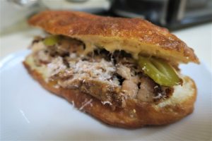 תקריב של כריך במילוי פרוסות בשר, חמוצים וגבינה מגוררת, מוגש בלחמניית בייגלה. הכריך על צלחת לבנה, עם רקע מטושטש המכיל פריטי מטבח.