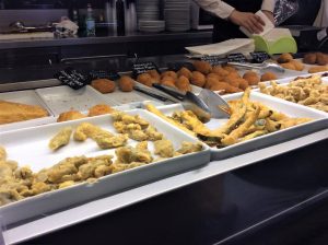 תצוגה של מאכלים מטוגנים שונים על מגשים לבנים בדלפק אוכל. המגשים מכילים פריטים כמו כדורי לחמים וירקות חבוטים. מלקחיים מונחים על אחד המגשים. כמה שלטים בכתב יד מציינים מנות שונות.