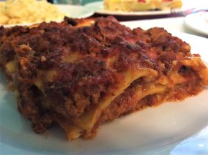 פרוסת לזניה עם שכבות גלויות של פסטה, רוטב בשר וגבינה נמצאת על צלחת לבנה. ברקע נראים צלחות אוכל מטושטשות, כולל מנה שנראית כפסטה או קדירה. הפוקוס הוא על פרוסת הלזניה.