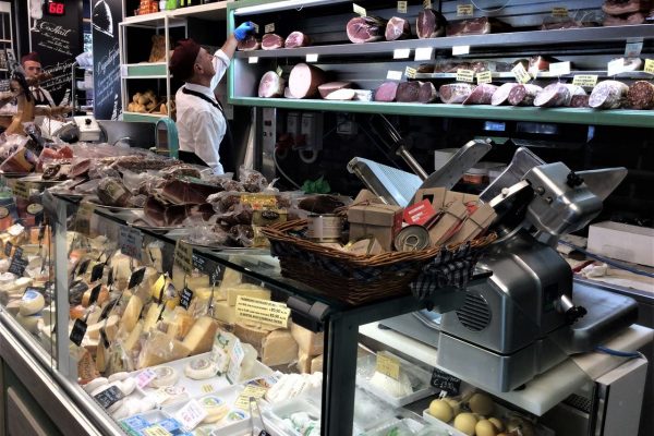 דלפק מעדניות המציג גבינות שונות, בשרים ומוצרים ארוזים. עובד במדים וכובע מושיט יד כדי להתאים פריטים במדף העליון. מכונת חיתוך נראית על השיש. הסצנה הומה מוצרים ופעילות.