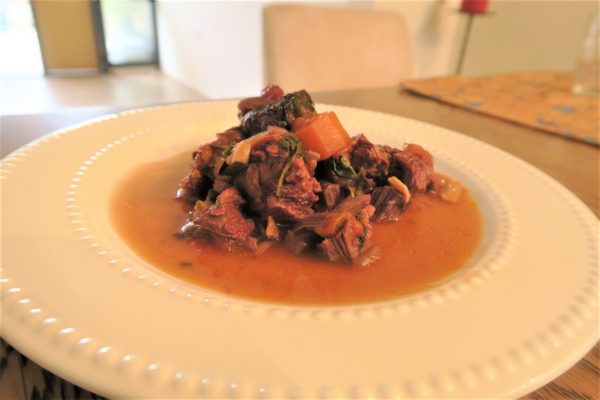 צלחת לבנה מכילה תבשיל עם נתחי בשר, גזר ועלים ירוקים במרק עשיר. ברקע נראה פנים מטושטש עם שולחן ונר אדום מרחוק.