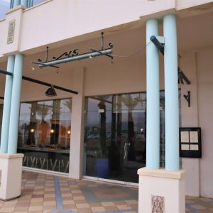 מראה חיצוני של בניין הכולל חלונות זכוכית גדולים ועמודים תכלת. לפטיו המרוצף דוגמה גיאומטרית. שני שלטים ריקים מותקנים ליד הכניסה. בפנים נראים אורות תלויים ושולחנות.