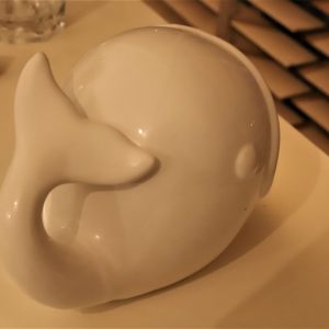 צלמית לוויתן קטנה ולבנה מונחת על צידו על משטח בהיר. ברקע יש פקק וזכוכית יחד עם כמה תריסי עץ.