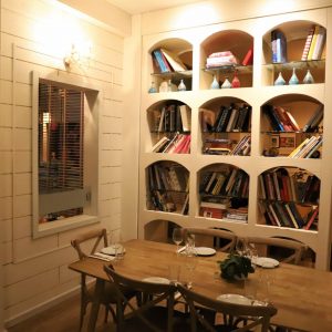 פינת אוכל נעימה כוללת שולחן עץ ערוך עם כוסות וצלחות, מוקף בכיסאות עץ. מדף ספרים גדול מלא בספרים ופריטי נוי עומד על הקיר. תאורה חמה ותמונות ממוסגרות נראות ברקע.