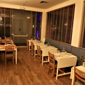 מסעדה קטנה וריקה עם ריצוף עץ ושולחנות ערוכים לסעודה. בחדר יש כיסאות עץ, מפות לבנות ותריסים המכסים את החלונות. התאורה רכה וחמימה, משרה אווירה נעימה.