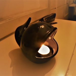 נר קטן דולק מונח בתוך מחזיק קרמיקה שחור בצורת לוויתן. המחזיק מונח על משטח בהיר ליד קיר. אור הנר מטיל זוהר חם על האזור שמסביב.