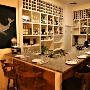 בתמונה בר עץ בצורת U עם כיסאות גבוהים. הבר משובץ בצלחות, סכו