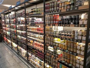 מדפים בחנות מציגים מגוון רחב של מוצרי מזון משומרים וקנקנים. המדפים מאורגנים היטב ומצוידים במלואם בתוויות ואריזות שונות, היוצרים סידור צבעוני. לחנות רצפת שיש.