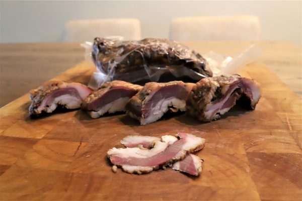 פרוסות בשר מבושלות מונחות על קרש חיתוך מעץ. מאחורי הפרוסות יש אריזה סגורה בוואקום עם עוד מהבשר. המשטח עשוי עץ, המספק תחושה כפרית למצגת.