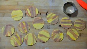 עוגיות שיש עגולות ולא מבושלות מסודרות בשורות על משטח עץ. חותכן עוגיות מתכת וסכין אדומה קטנה מונחים בקרבת מקום. לעוגיות יש דוגמה של מערבולות צהובות וחומות.