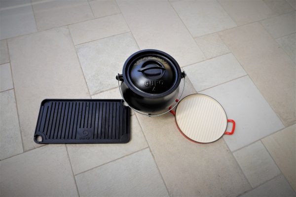 סיר ברזל יצוק שחור עם מכסה וידית מונח על רצפת אריחים בהירה. לצידו שתי מחבתות: האחת מלבנית עם קווי גריל, והשנייה עגולה, לבנה עם שפה אדומה, בעלת משטח חלק.