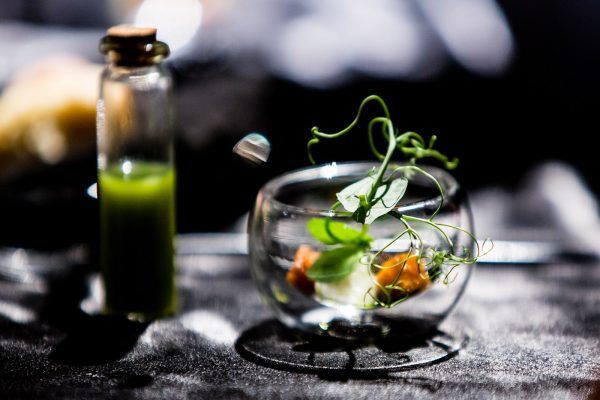קערת זכוכית מכילה מנה קטנה ומוצגת באומנות עם ירקות וחתיכות אוכל קטנות. בקבוק זכוכית קטן מלא בנוזל ירוק נמצא ליד הקערה. הרקע מטושטש וכהה.