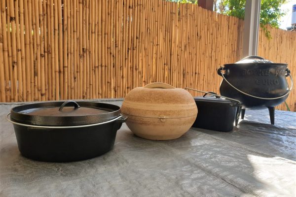 אוסף של ארבעה סירי בישול על משטח אפור. משמאל לימין: סיר ברזל יצוק שחור עם מכסה, סיר חרס עגול, סיר מלבני שחור וסיר שחור עם רגליים ומכסה. גדר במבוק נמצאת ברקע.