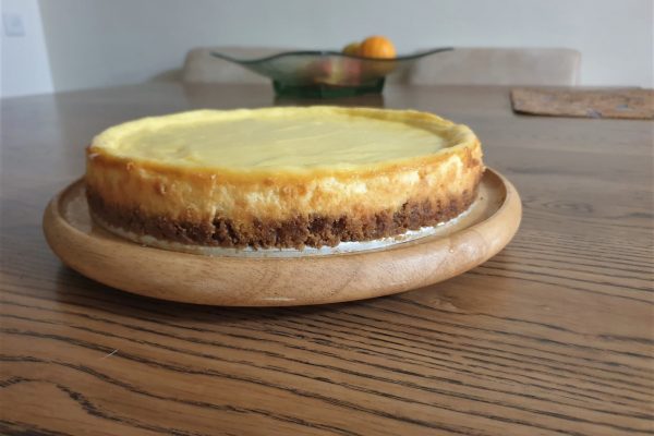 עוגת גבינה עם פלטה מוזהבת יושבת על צלחת עץ עגולה על שולחן עץ. ברקע ניצבת קערת זכוכית עם פירות, כולל תפוז, המונחת על שולחן אוכל.