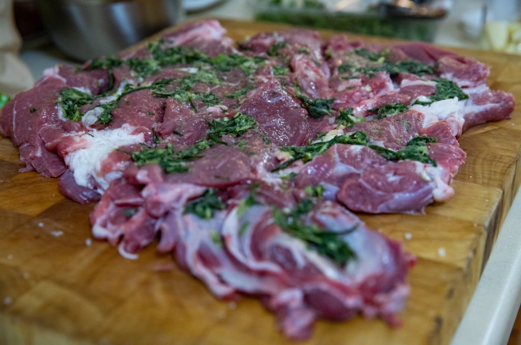 בשר טלה נא פרוס על קרש חיתוך עץ, ומעליו עשבי תיבול קצוצים. הבשר נראה משויש בשומן ומוכן לבישול. רקע מטושטש מציג כלי מטבח ומרכיבים.