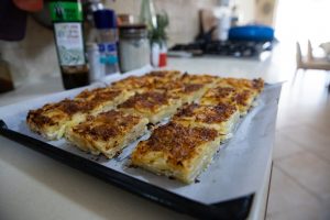 תבנית אפייה מרופדת בנייר אפייה מונחת על משטח עבודה במטבח, ועליה שישה־עשר חתיכות שוות בגודל של מאפה תפוחי אדמה אפוי בשכבות. ברקע נראים תבלינים וחפצים נוספים מהמטבח.