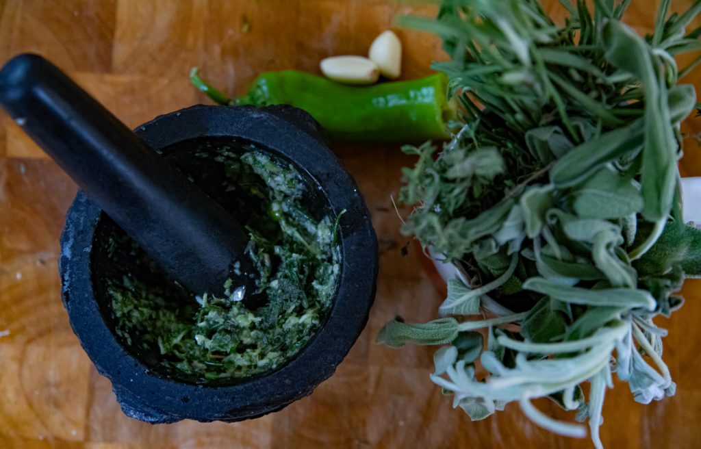 מכתש ועלי עם משחה ירוקה לצד עשבי תיבול טריים, שיני שום ופלפל צ'ילי ירוק על משטח עץ.