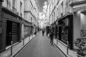 סצנת רחוב צרה בשחור-לבן עם בניינים גבוהים משני הצדדים. אנשים הולכים לאורך המדרכה. חנויות עם שלטים וחלונות ראווה לאורך הרחוב. שמים מעוננים חלקית נראים מעל.