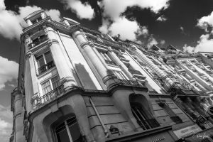 תמונה בשחור-לבן של בניין אירופאי קלאסי עם ארכיטקטורה מקושטת במבט מזווית נמוכה. החזית כוללת מרפסות ואלמנטים דקורטיביים. עננים וצללים נראים בשמים מעל.