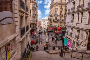 סצנת רחוב בפריז מציגה גרם מדרגות יורד המוביל לשדרה הומה. אנשים מסתובבים בין חנויות, עם שלט כניסה אדום למטרו גלוי. בניינים גבוהים ומעוטרים לאורך הרחוב תחת שמים מעוננים חלקית.