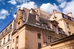 בניין אבן בז' ואפור עם גגות צפחה תלולים ומספר ארובות ניצב מתחת לשמים כחולים עם עננים מפוזרים. המבנה כולל חלונות שונים ופרטים אדריכליים בסגנון אירופאי קלאסי.