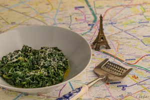 קערת תרד שמנת עם גבינה מגוררת מונחת על המפה. מגרדת גבינה עם ידית כחולה פרחונית שוכנת בסמוך. צלמית קטנה של מגדל אייפל עומדת ברקע.