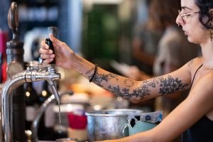 אדם עם קעקועים פרחוניים על זרועו ממלא כוס מים מברז. הם מרכיבים משקפיים ונראה שהם נמצאים בסביבה של בר או בית קפה, עם בקבוקים ברקע.