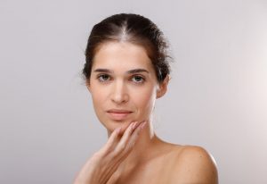 אישה ששיערה קשור לאחור נוגעת בעדינות בסנטרה בידה הימנית. יש לה הבעה ניטרלית ועור פנים חלק. הרקע פשוט ובהיר, מבליט את תווי פניה.