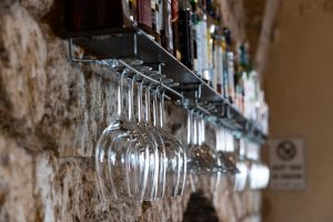 שורה של כוסות יין ריקות תלויות הפוך מתחת לבקבוקים על מתלה מתכת המחובר לקיר אבן. שלט קטן עם טקסט בלתי קריא נראה מימין. נראה כי התפאורה היא בר כפרי או מרתף יין.