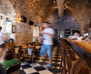 פנים מסעדה כפרי עם קירות אבן וריצוף משובץ. אדם בתנועה לבוש בחולצה לבנה ומכנסיים קצרים כחולים חולף על פניו ויוצר אפקט טשטוש. הלקוחות יושבים ליד השולחנות. הקיר האחורי כולל אמנות דקורטיבית של לוח גיר.